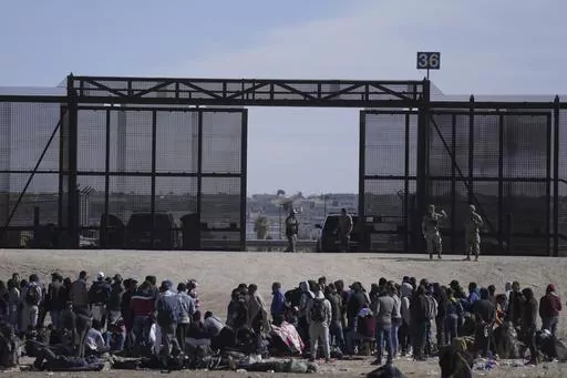 Migrants who crossed the border from Mexico into the U.S. wait next to the U.S. border wall where U.S. Border Patrol agents stand guard, seen from Ciudad Juarez, Mexico, Thursday, March 30, 2023. The Biden administration will open migration centers in South and Central America for asylum seekers heading to the U.S.-Mexico border, in a bid to slow what’s expected to be a surge of migrants seeking to cross the border next month as pandemic-era immigration restrictions end, U.S. officials said Th