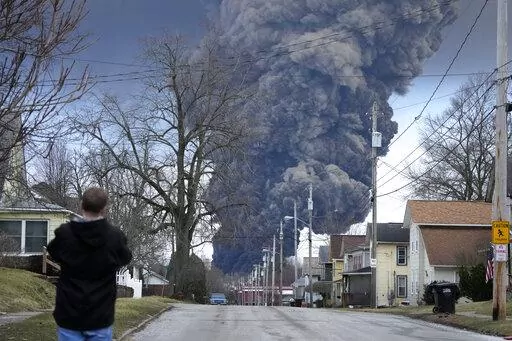 A man takes photos as a black plume rises over East Palestine, Ohio, as a result of a controlled detonation of a portion of the derailed Norfolk Southern train, Feb. 6, 2023. After toxic chemicals were released into the air from a wrecked train in Ohio, evacuated residents remain in the dark about what toxic substances are lingering in their vacated neighborhoods while they await approval to return home. (AP Photo/Gene J. Puskar, File)