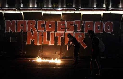 Demonstrators start a fire at the foot of the barrier protecting the National Palace during a march to commemorate the 9th anniversary of the disappearance of 43 missing Ayotzinapa university students, in Mexico City, Tuesday, Sept. 26, 2023. The graffiti on the barrier reads "Military Narcostate" in Spanish. (AP Photo/Marco Ugarte)