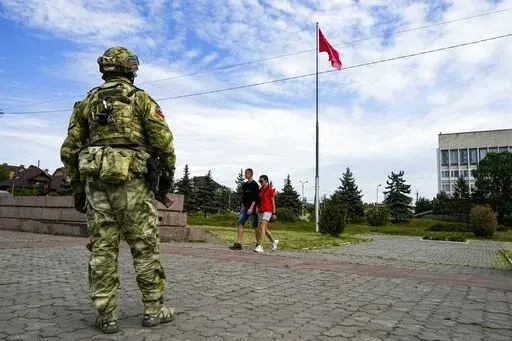 A young couple walks past a Russian soldier guarding an area at the Alley of Glory exploits of the heroes - natives of the Kherson region, who took part in the liberation of the region from the Nazi invaders, in Kherson, Kherson region, south Ukraine, Friday, May 20, 2022, with a replica of the Victory banner marking the 77th anniversary of the end of World War II right in the background. Ukrainian forces pressing an offensive in the south have zeroed in on Kherson, a provincial capital that has