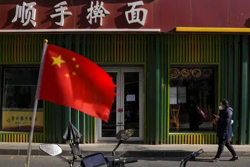 A woman wearing a face mask walks past a national flag outside a closed noodles restaurant due to the COVID-19 controls in Beijing, Wednesday, Nov. 23, 2022. (AP Photo/Andy Wong, File)