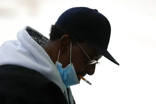 A man with a protective mask smokes a cigarette while waiting for a bus in Detroit, Wednesday, April 8, 2020. U.S. cigarette smoking dropped to a new all-time low in 2020, with 1 in 8 adults saying they were current smokers, according to survey data released Thursday, March 17, 2022, by the Centers for Disease Control and Prevention. Adult e-cigarette use also dropped, the CDC reported. (AP Photo/Paul Sancya, File)