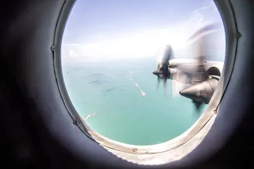 In this photo provided by the New Zealand Defense Force, an Orion plane flies over Tonga where volcanic ash covers roof tops and vegetation, Monday, Jan. 17, 2022. Thick ash on an airport runway was delaying aid deliveries to the Pacific island nation of Tonga, where significant damage was being reported days after a huge undersea volcanic eruption and tsunami. (CPL Vanessa Parker/NZDF via AP)