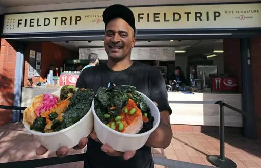 This Aug. 30, 2019 photo shows chef JJ Johnson posing with two signature rice bowls, one salmon and one vegetable, outside his Field Trip counter-service restaurant kiosk on the food court at the US Open tennis championships in New York. Johnson's new cookbook "The Simple Art of Rice: Recipes from Around the World for the Heart of Your Table," a book co-authored with Danica Novgorodoff, celebrates rice's versatility. (AP Photo/Charles Krupa, File)