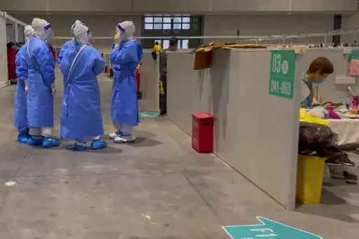 In this image taken from video provided by Beibei, who asked to be identified only by her given name, medical workers wearing protective suits chat as a resident takes a rest at the National Exhibition and Convention Center on April 15, 2022, in Shanghai.  The convention center converted to a quarantine facility set up for people who tested positive but have few or no symptoms. (Beibei via AP)