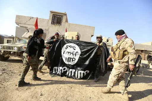Iraqi Army soldiers celebrate as they hold a flag of the Islamic State group they captured during a military operation to regain control of a village outside Mosul, Iraq, Nov. 29, 2016. Ten years after the Islamic State group declared its caliphate in large parts of Iraq and Syria, the extremists now control no land, have lost many prominent founding leaders and are mostly away from the world news headlines. (AP Photo/Hadi Mizban, File)