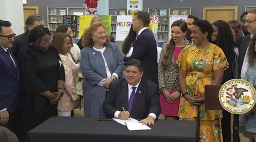 In this screenshot from a livestream broadcast by the State of Illinois, Gov. J.B. Pritzker signs a bill, Monday, June 12, 2023, at Harold Washington Library's Thomas Hughes Children's Library in downtown Chicago. The new law will require the state's libraries to uphold a pledge not to ban material because of partisan disapproval, starting on Jan. 1, 2024. If they refuse, they will not receive state funding. Pritzker said the law will make Illinois the first state in the nation to outlaw book ba